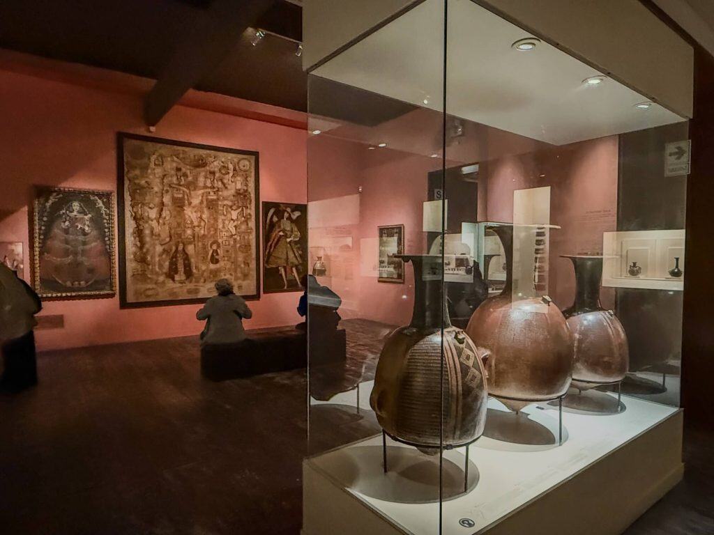 A lowly lit room with red walls, paintings, and encased artifacts of black and brown large pottery with painting designs on the exterior.
