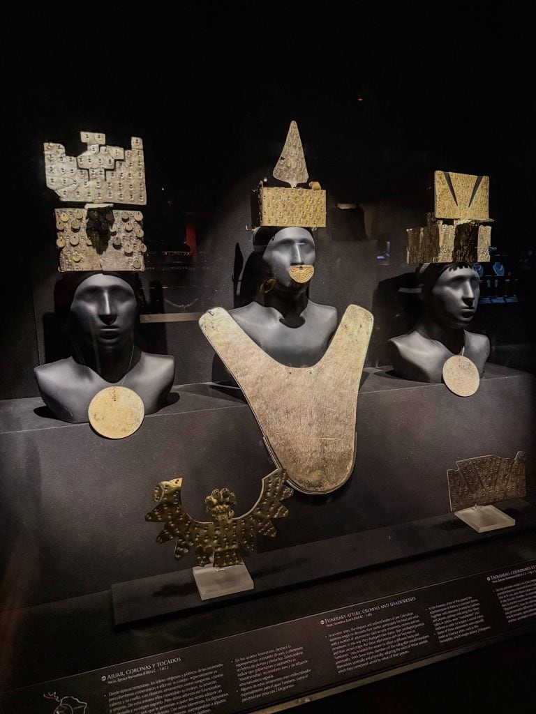 Three black mannequin heads wearing large gold headpieces and neck plates that are ancient pieces to see on display at Larco Museum.