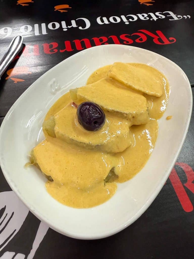 A white plate with thick slices of steamed potato covered in a yellow sauce and topped with an olive at a food stall in Mercado Surquillo.