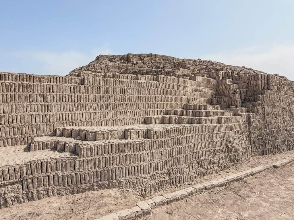 A side view of the Huaca Pucllana pyramid ruins made of clay and adobe bricks stacked in a vertical orientation with a series of steps or levels exposed to see.