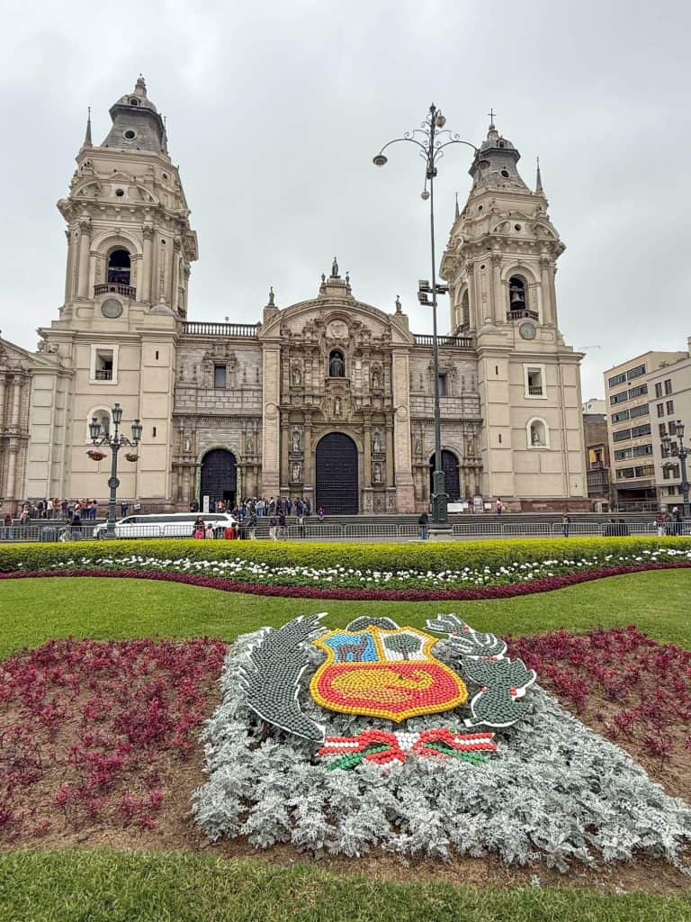 The exterior view of the Lima Cathedral with its colonial style of architecture with a patch of manicured grass and flowers in the foreground of the square in Lima's historic district.