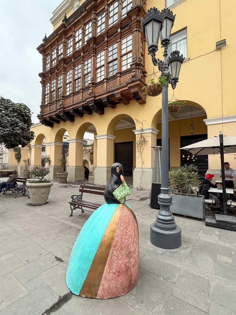 A statue of a woman in a dress with her head and face covered in a veil with only one eye showing that are all over the historic center of Lima, Peru with a building behind that has enclosed wooden box balconies.