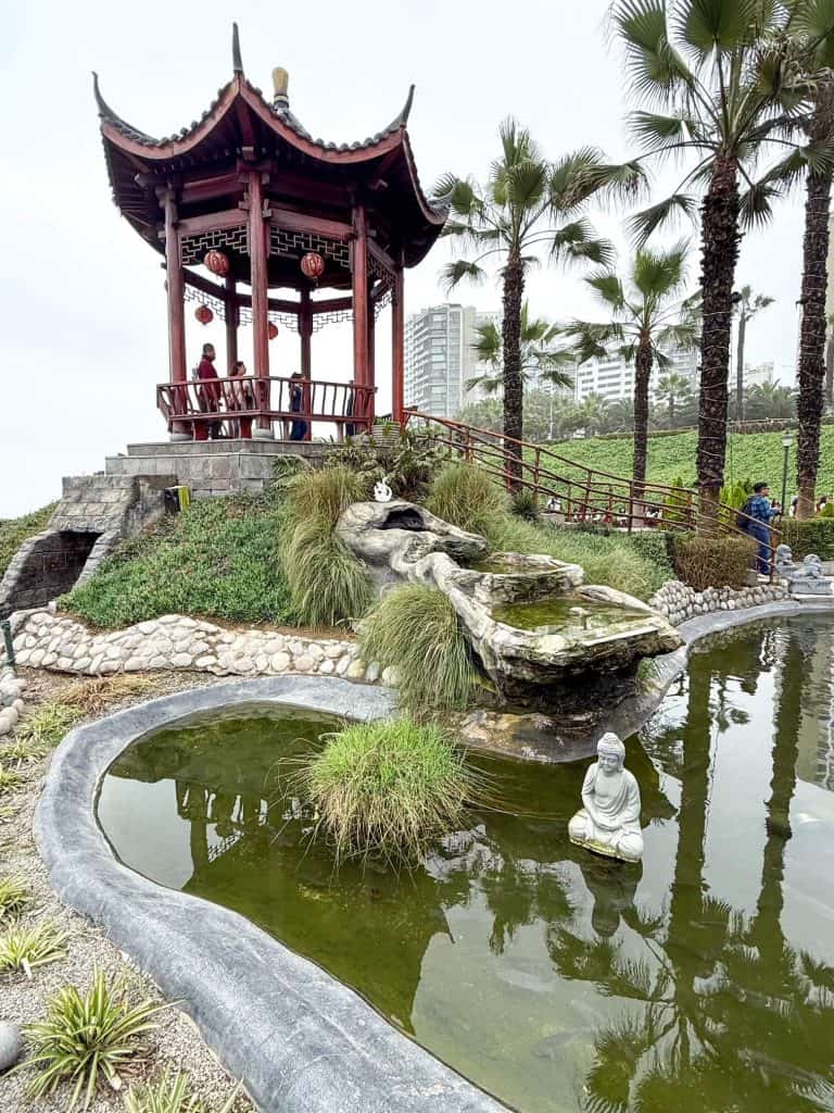 A small coy pond with a buddha statue below a Chinese gazebo surrounded with pam trees at Parque Chino along the coast in Miraflores, Lima.