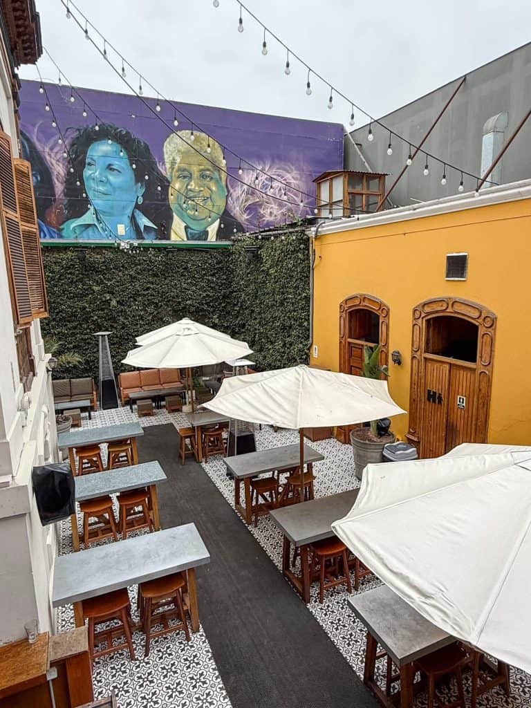 Looking down at an outer courtyard of a restaurant in Barranco with white umbrella tables, hanging lights, and a colorful art mural of 3 people on the back wall in Lima, Peru.