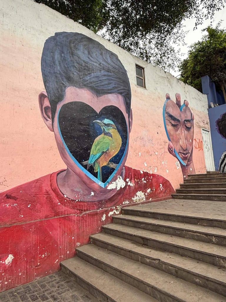 A vibrant art mural of a boy with his face cut out in the shape of a heart and him holding his heart face shaped face in his hand in red, orange, and green colors in Lima's artsy district.