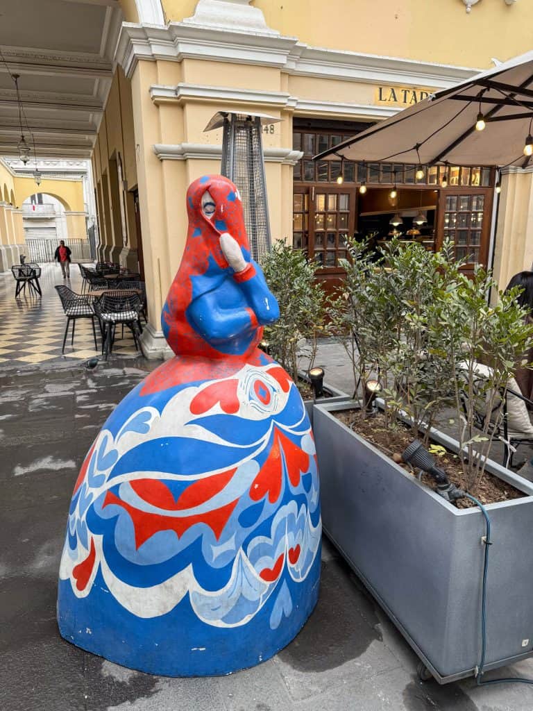A colorful sculpture of a woman in a square in Lima's historic district with a red, white, and blue painted dress and a shawl covering her face except for one eye in front of a cafe.